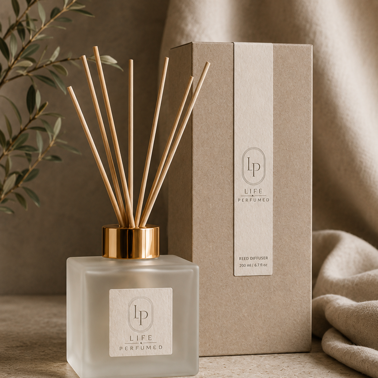 A Life Perfumed reed diffuser bottle with natural wood reeds and a gold collar, shown in front of its kraft paper box on a neutral beige surface with draped fabric and a leafy branch in the background.